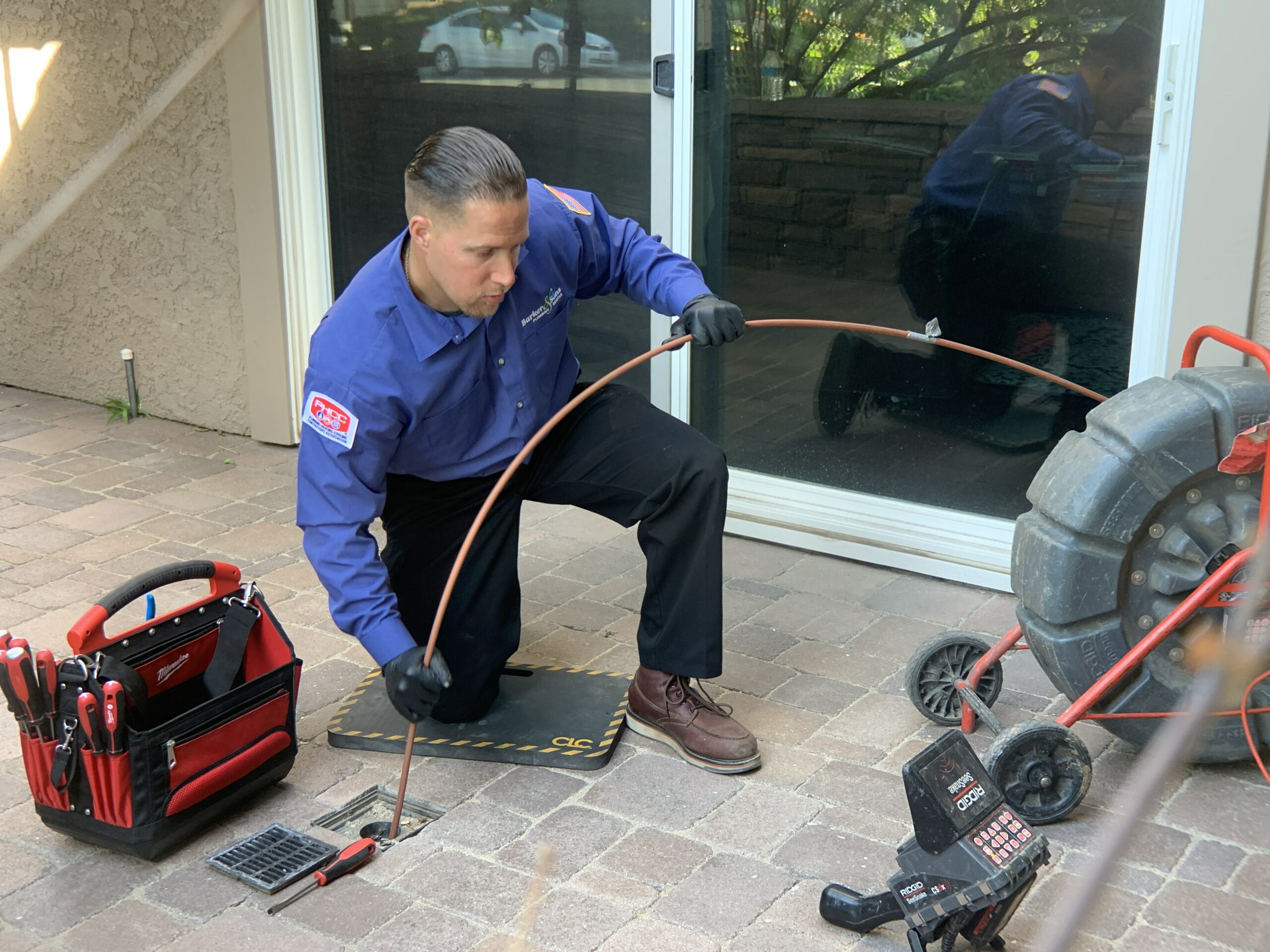 A plumber in a blue shirt kneels on a mat and skillfully uses a tool to clear the drain outside the house. His trusty tool bag and equipment sit on a cart nearby, ready for any challenge.