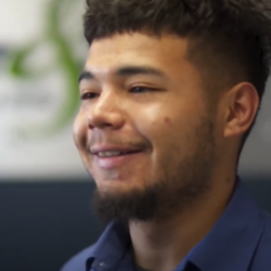 A friendly plumber with short curly hair and a beard smiles slightly in a blurred indoor setting, ready to tackle any plumbing challenge while wearing a blue shirt.