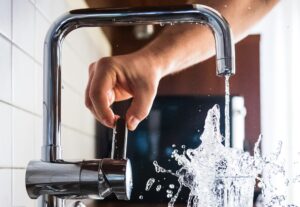 Stainless steel sink faucet turned on. Water splashes into a glass cup.