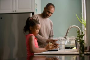 A man and a girl stand at the kitchen sink, washing dishes to keep the water bill low. She holds a dish while he rinses another, both mindful of water use. A dish rack and green plant sit on the counter nearby.