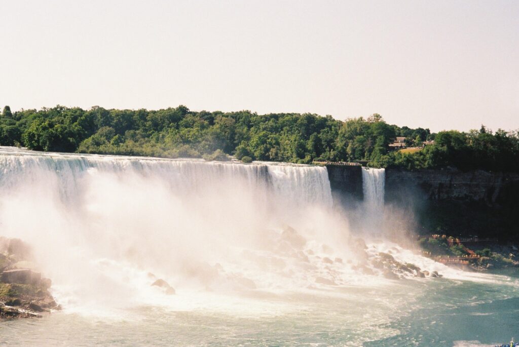 A large waterfall cascades with breathtaking force, sending mist rising gracefully into the air amidst lush greenery. A building stands in the distance under a clear sky, quietly observing the powerful display.
