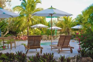 Two wooden lounge chairs face a small round pool, surrounded by white umbrellas, tropical plants, and palm trees. This inviting outdoor retreat offers a perfect spot to relax during an Anaheim heat wave, while smart landscaping shields the plumbing from the sun.