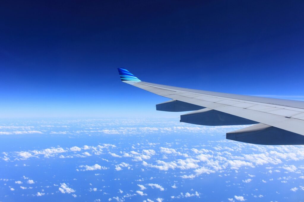 An airplane wing soars above a blanket of clouds, framed by a clear blue sky and the distant horizon—a perfect reminder to review plumbing tips and safeguard your home’s pipes before leaving for your summer vacation.