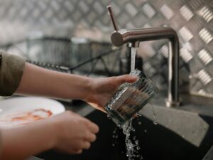 A person washes a clear glass under running water at the kitchen sink, scrubbing to remove hard water stains. A dish rack and plate sit in the background.