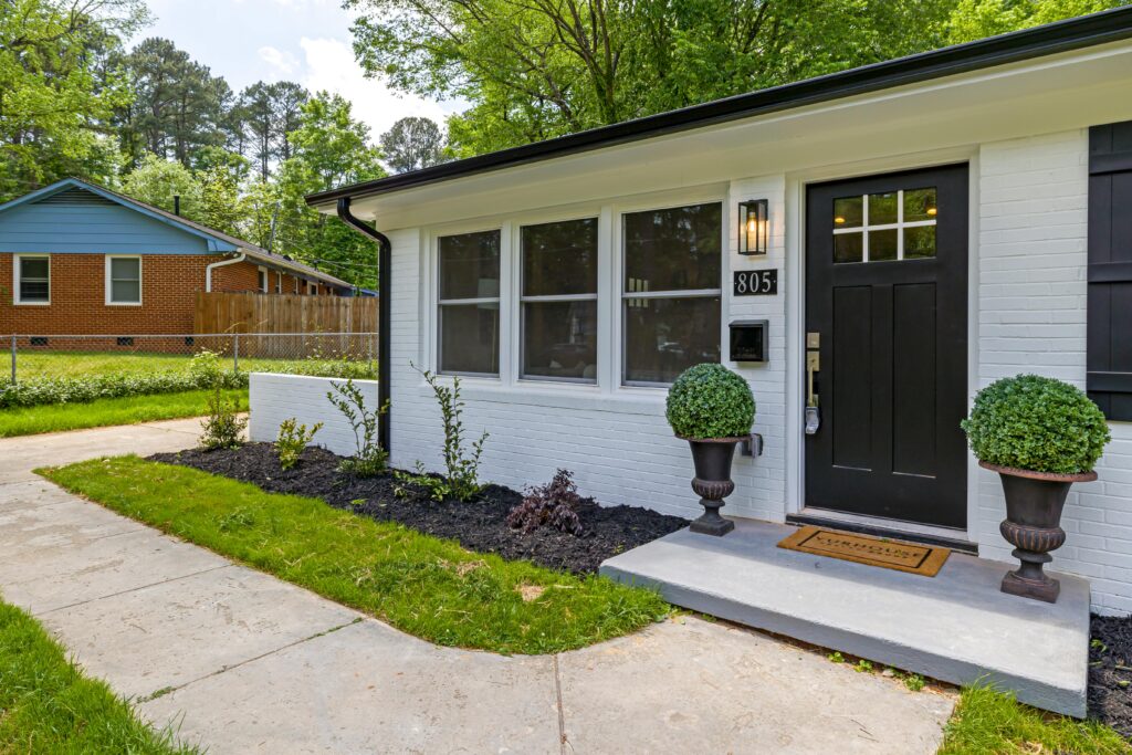 A white brick home with black trim and a black door greets visitors. Two potted shrubs and a welcome mat sit by the entrance, next to a sidewalk and landscaped garden in Orange County.