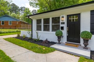 A white brick home with black trim and a black door greets visitors. Two potted shrubs and a welcome mat sit by the entrance, next to a sidewalk and landscaped garden in Orange County.