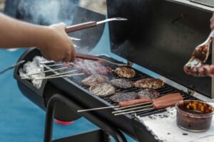 Grill burgers, hot dogs, and meat skewers on a barbecue for Memorial Day. Smoke rises from the grill as a bowl of sauce sits nearby.