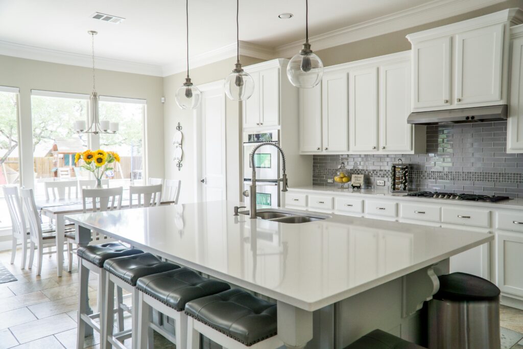 Upgrade your kitchen with modern white cabinets, a spacious island paired with gray stools, and sleek stainless steel appliances. Brighten the space with pendant lights, and set a dining table decorated with sunflowers near expansive windows—ideal for summer hosting.