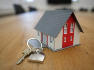 A small model house with a red facade stands on a wooden table beside a set of keys, capturing the excitement and key milestones, like home inspection, involved in buying a home.