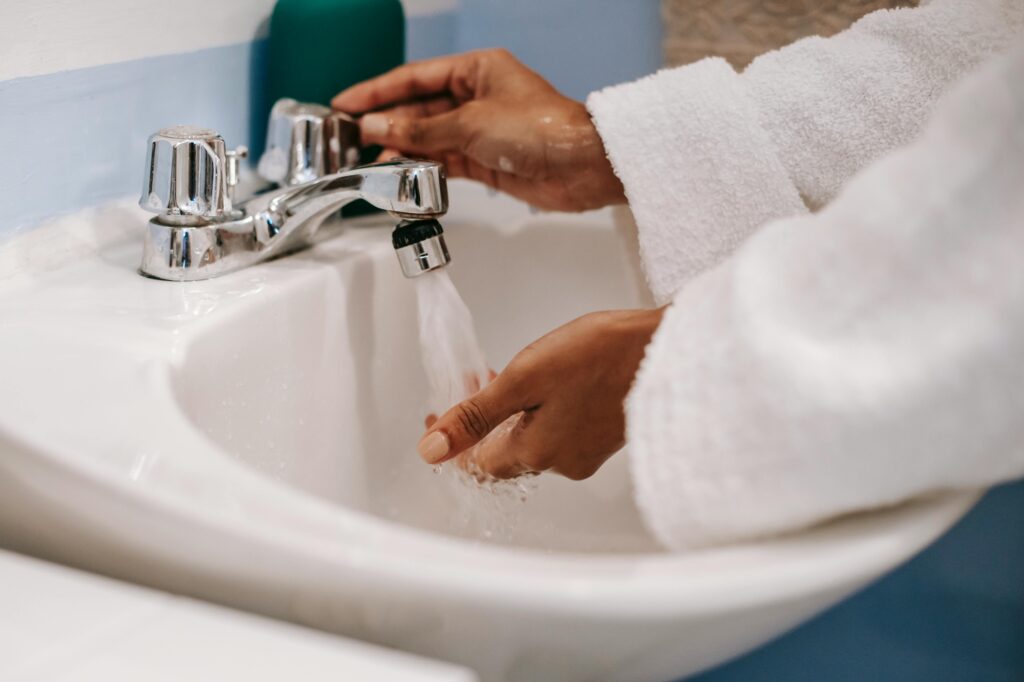Wearing a white robe, a person washes their hands under running water at a bathroom sink with chrome faucets, demonstrating the efficiency of an upgraded Orange County plumbing system.