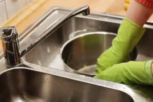 A person in green gloves washes a metal pot in a stainless steel sink under the faucet—an ideal image for any summer plumbing guide or a comprehensive kitchen upkeep manual.