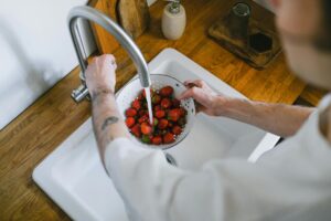 Someone rinses strawberries in a metal colander under running water, making sure no food debris or other items clog the kitchen sink drain.