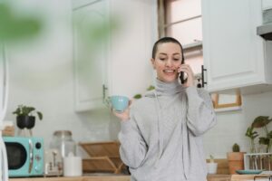 A person with short hair, wearing a gray hoodie, stands in the kitchen holding a blue mug and calls an emergency plumber for urgent help.
