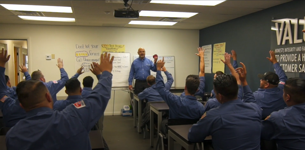 A man leads a classroom as students in blue uniforms raise their hands. Notes, charts cover the walls and a projector hangs above.