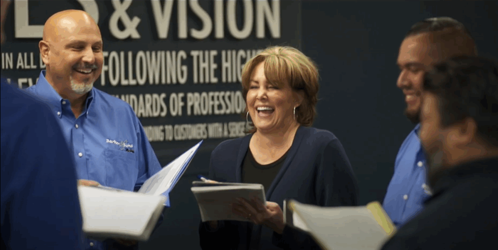 Four adults gather in front of a wall sign reading "VISION" and other text, holding notepads and smiling at the camera.