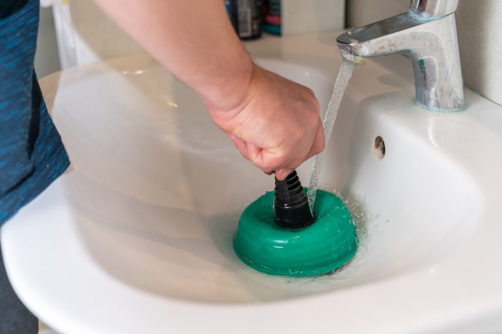 A person unclogs a bathroom sink with a green plunger as water runs from the faucet, showing effective DIY drain cleaning.