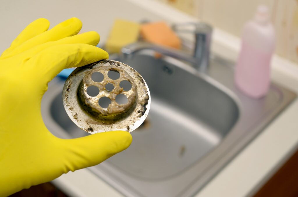 A yellow-gloved hand holds a dirty metal sink drain cover above a kitchen sink; cleaning supplies sit in the background.