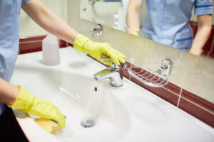 Wearing yellow gloves, a person cleans a bathroom sink, runs water, uses a sponge, and adjusts the tap to prevent plumbing issues.