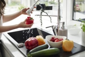 A person washes a red bell pepper under a kitchen faucet as a camera captures clean prep spaces and tips to prevent clogged drains.