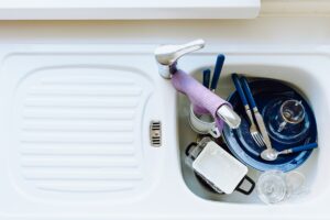 A white kitchen sink holds unwashed dishes, utensils, and cups; a purple towel drapes over the faucet, showing drain differences.
