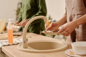 Two people stand at a kitchen sink. One washes dishes as the other prepares food. A soap dispenser on the counter highlights plumbing care.